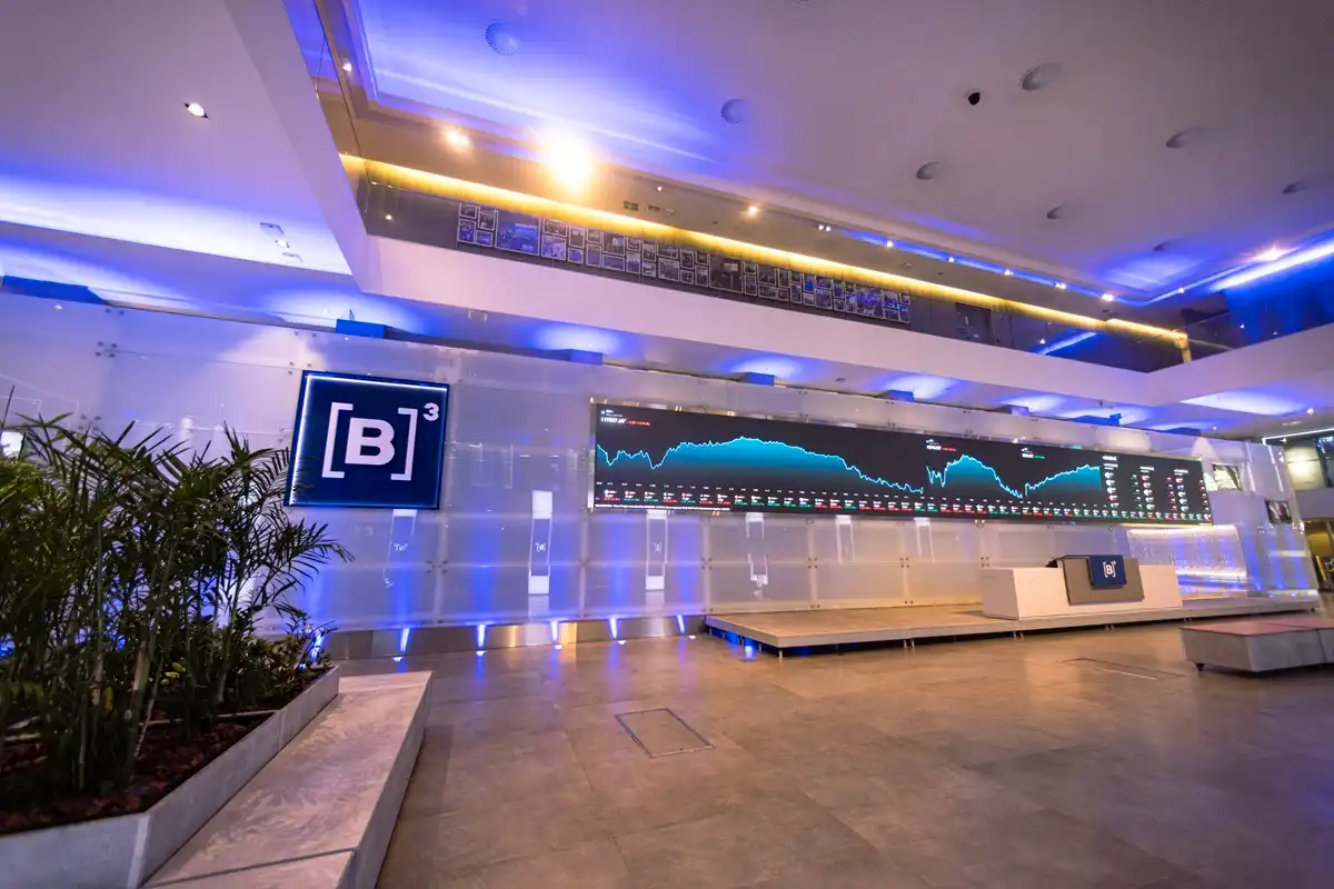Painel principal da B3 em São Paulo, com logotipo da Bolsa e gráfico digital exibindo variação do mercado financeiro em ambiente iluminado em tons de azul.