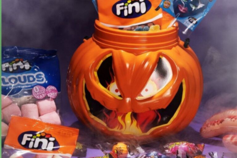 Balde em formato de abóbora de Halloween cheio de doces Fini, com embalagens coloridas e produtos como marshmallows e balas sortidas, sobre fundo roxo com efeito de fumaça.
