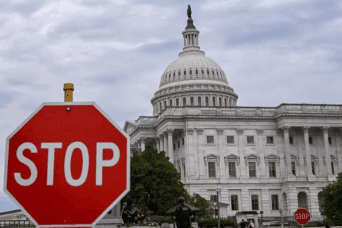Placa de trânsito com a palavra STOP em frente ao prédio do Capitólio dos Estados Unidos em Washington, simbolizando a paralisação do governo (shutdown).