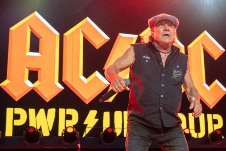 O vocalista da banda AC/DC, Brian Johnson, no palco, segurando um microfone. Ele usa um boné tipo 'boina' cinza, uma camisa preta sem mangas e calças escuras. Atrás dele, o grande logotipo laranja da banda AC/DC e a inscrição 'PWR UP' iluminada.