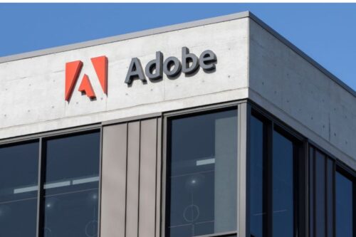 O logotipo da Adobe, com um 'A' estilizado em vermelho e o nome 'Adobe' em preto, está montado no topo de um edifício de escritórios moderno de concreto sob um céu azul claro.