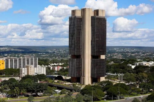 Vista externa do Edifício-Sede do Banco Central do Brasil (BCB) em Brasília, com sua arquitetura brutalista característica, torres escuras verticais e detalhes horizontais claros, sob um céu azul com nuvens brancas, rodeado por uma vasta área verde e a paisagem urbana da capital federal ao fundo.