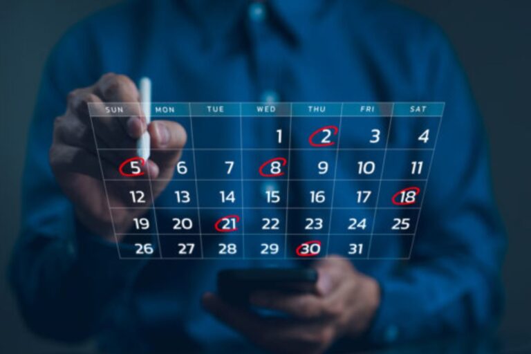 Uma pessoa, em um ambiente de luz azul, está marcando a data '5' (domingo) em um calendário digital flutuante com uma caneta stylus. As datas '2', '5', '8', '18', '21' e '30' estão circuladas, sugerindo o agendamento de eventos ou compromissos importantes ao longo do mês.