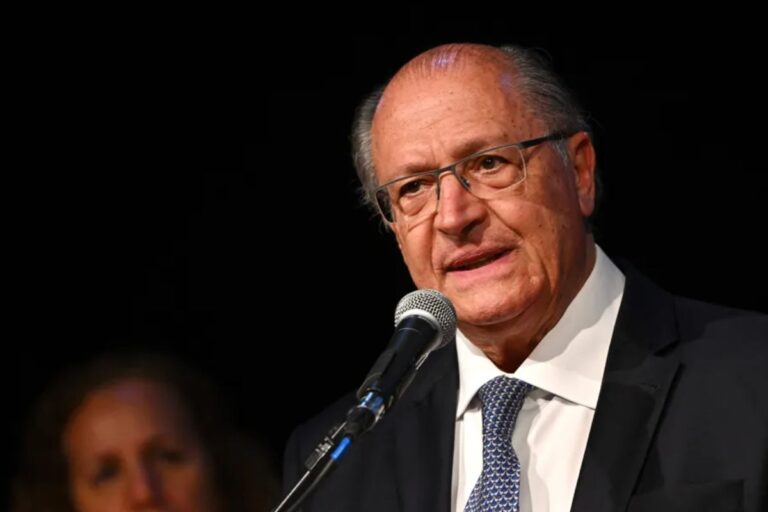 Geraldo Alckmin, político brasileiro, em close-up, discursando em um microfone com fundo escuro. Ele veste um terno e gravata, e usa óculos.