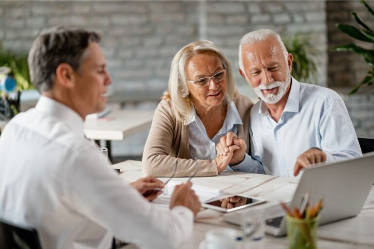 Um casal de idosos, sorrindo e de mãos dadas, sentado em uma mesa de escritório enquanto revisam documentos e apontam para a tela de um laptop, com um consultor financeiro ao lado esquerdo.