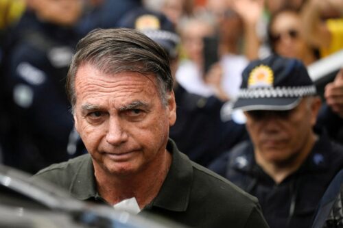 Foto em close-up de Jair Bolsonaro. Ele está em primeiro plano, com uma expressão séria, vestindo uma camisa polo verde-escura e apresenta um curativo branco na base do pescoço.