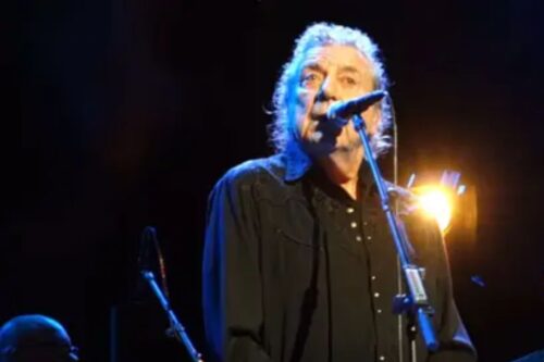 O cantor Robert Plant, do Led Zeppelin, em close-up no palco, cantando em um microfone. Ele tem cabelos grisalhos e longos, veste uma camisa preta e está sob luzes de show, com uma forte luz amarela iluminando a cena por trás. A imagem é de uma apresentação histórica do clássico 'Kashmir' em Londres.