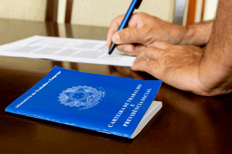 Pessoa assinando a Carteira de Trabalho e Previdência Social ao lado de um documento, possivelmente um formulário ou papel de registros.