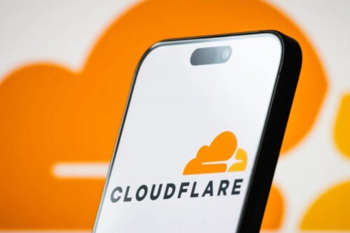 O logo da Cloudflare, um ícone de nuvem estilizada em laranja e amarelo, é exibido na tela de um smartphone. O fundo é um desfoque de cor laranja com o mesmo formato de nuvem. Esta imagem ilustra a interrupção de serviço da Cloudflare que afetou plataformas globais.