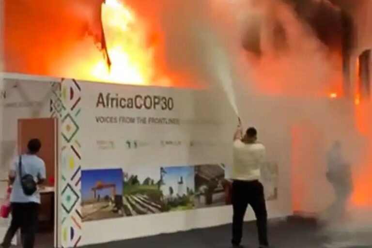 Uma cena de incêndio, onde chamas intensas e fumaça laranja enchem a parte superior do quadro. Um homem no centro está usando um extintor de incêndio para combater o fogo que atinge uma parede ao lado de um estande de exibição com o banner 'AfricaCOP30: Voices from the Frontlines'.
