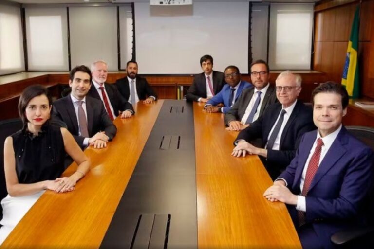 Membros do Comitê de Política Monetária (Copom) do Banco Central do Brasil sentados ao redor de uma longa mesa de reunião em uma sala formal. O grupo é composto por nove pessoas, em sua maioria homens, vestindo trajes sociais. Uma bandeira do Brasil é visível à direita.