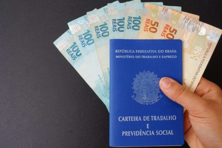 Uma mão segura a capa azul da Carteira de Trabalho e Previdência Social (CTPS) brasileira, colocada sobre uma pilha de notas de Real (R$ 50 e R$ 100), simbolizando trabalho, direitos e pagamento.