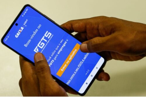 Close-up de duas mãos segurando um smartphone que exibe a tela de boas-vindas do aplicativo do FGTS (Fundo de Garantia do Tempo de Serviço), com o logotipo da Caixa e um botão em destaque escrito 'Entrar no aplicativo'.
