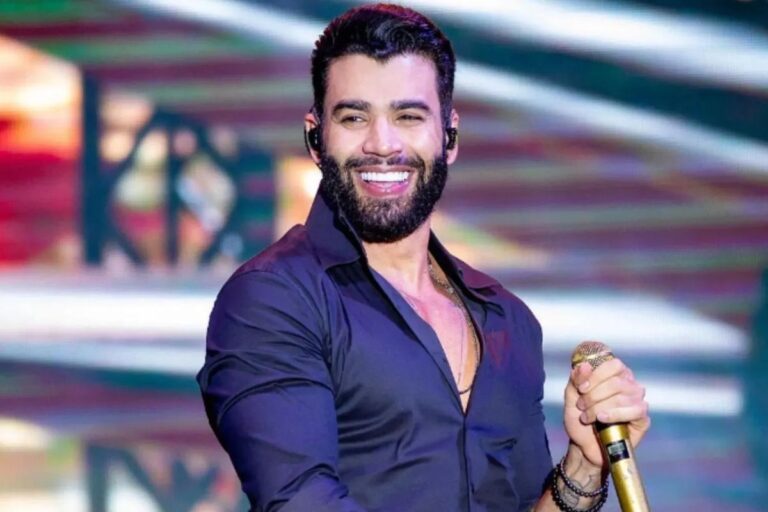 O cantor sertanejo Gusttavo Lima, de barba escura e camisa preta, sorrindo e olhando para frente durante uma apresentação no palco.