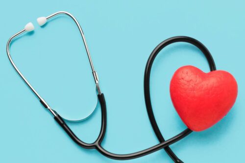 ista superior de um estetoscópio médico preto sobre um fundo azul claro. O tubo do estetoscópio faz uma curva suave, envolvendo um coração vermelho texturizado posicionado no lado direito da imagem.