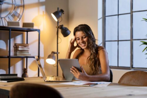 Mulher sorridente com cabelo castanho longo e óculos, sentada em uma mesa de madeira clara em um escritório em casa.