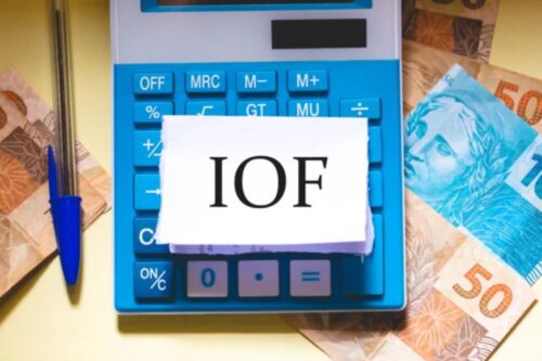 Uma calculadora azul e branca está sobre uma superfície amarela, cercada por notas de Real (moeda brasileira) de R$ 50 e R$ 100.