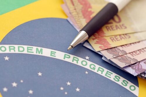 Uma imagem em close-up mostrando a faixa 'ORDEM E PROGRESSO' da bandeira do Brasil. Sobre a bandeira, estão algumas notas de Real brasileiro e uma caneta preta e prateada, com a ponta apontando para a faixa.