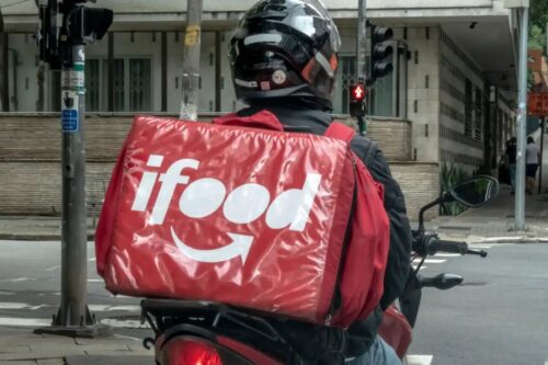 Entregador de comida delivery da iFood de costas, usando capacete e mochila térmica vermelha com logotipo da iFood, aguardando na rua em um pacote de cidade.