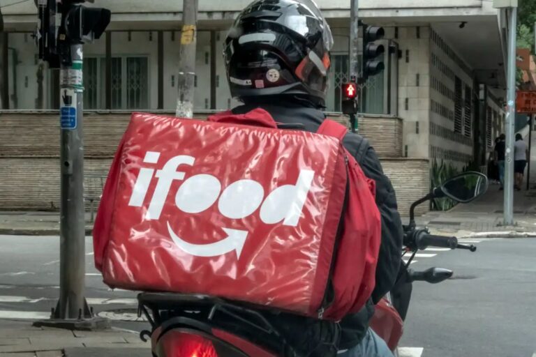 Entregador de comida delivery da iFood de costas, usando capacete e mochila térmica vermelha com logotipo da iFood, aguardando na rua em um pacote de cidade.