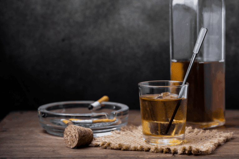 Cigarros, cinzeiro com pontas apagadas, bebida alcoólica em copo com canudo e garrafa de aguardente na mesa, ambiente com fundo escuro