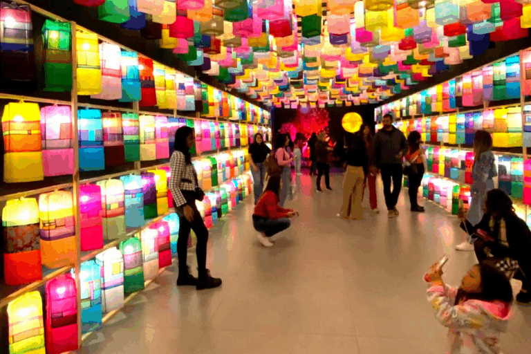 Pessoas explorando uma galeria de luzes decorativas coloridas, com paredes forradas de luminárias de diversos tons, criando um ambiente vibrante e encantador.