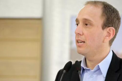 Rodrigo Manga vestindo um blazer escuro, falando em um evento. Ele está em um plano de médio-primeiro-plano, com dois microfones em frente e um fundo desfocado em tons de bege e branco.