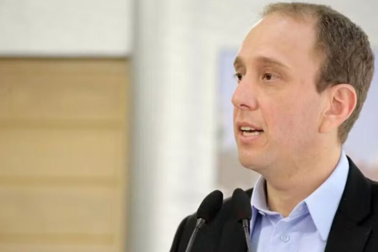 Rodrigo Manga vestindo um blazer escuro, falando em um evento. Ele está em um plano de médio-primeiro-plano, com dois microfones em frente e um fundo desfocado em tons de bege e branco.