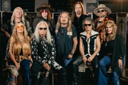 Foto de estúdio de grupo, em estilo vintage e rock and roll, com nove pessoas (seis homens e três mulheres) posando. Eles estão vestidos com roupas escuras e de couro, óculos escuros e chapéus, exibindo cabelos longos e barbas. A maioria está sentada ou agachada na frente.