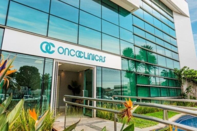 Fachada moderna de um edifício de consultórios ou clínica médica, com grandes janelas de vidro azul refletoras e a logo e nome "ONCOCLÍNICAS" em destaque na parte superior.
