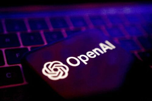 O logotipo e o nome 'OpenAI' em branco exibidos na tela escura de um celular ou dispositivo, posicionado sobre um teclado de computador com iluminação em tons de roxo e rosa.