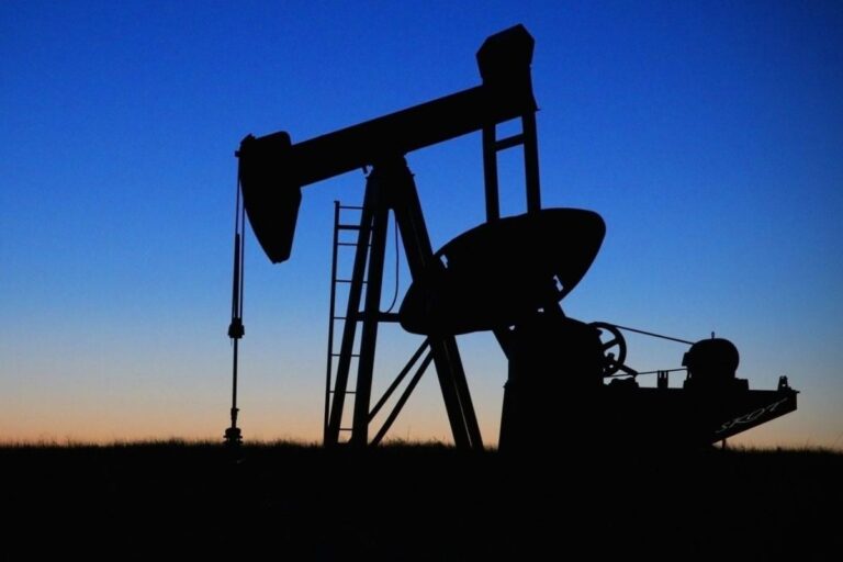 Silhueta de uma bomba de petróleo (cabeça de cavalo) contra um céu gradiente de pôr do sol/amanhecer, com tons de azul escuro e laranja fraco no horizonte.