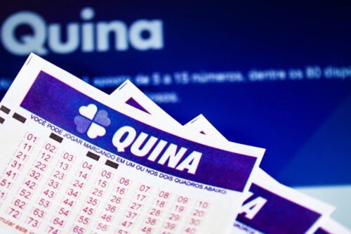 Bilhetes da loteria brasileira Quina, com o logotipo de trevo de quatro folhas e números para marcação, empilhados sobre um fundo azul escuro com a palavra "Quina" em destaque e embaçada.