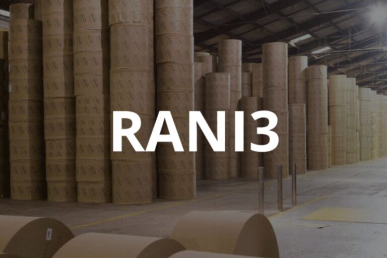 Abreviatura de ações RANI3 (Irani Papel e Embalagem) sobreposta à imagem de grandes pilhas de rolos de papelão ou celulose em um depósito industrial.