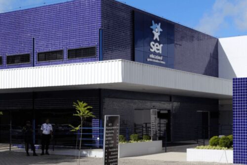 Fachada moderna do edifício da Ser Educacional. A parte superior é revestida com azulejos azuis e exibe o logotipo e o nome da empresa em branco. A entrada principal, com painéis escuros e um toldo branco horizontal, tem dois indivíduos uniformizados parados à esquerda e um jardim lateral.