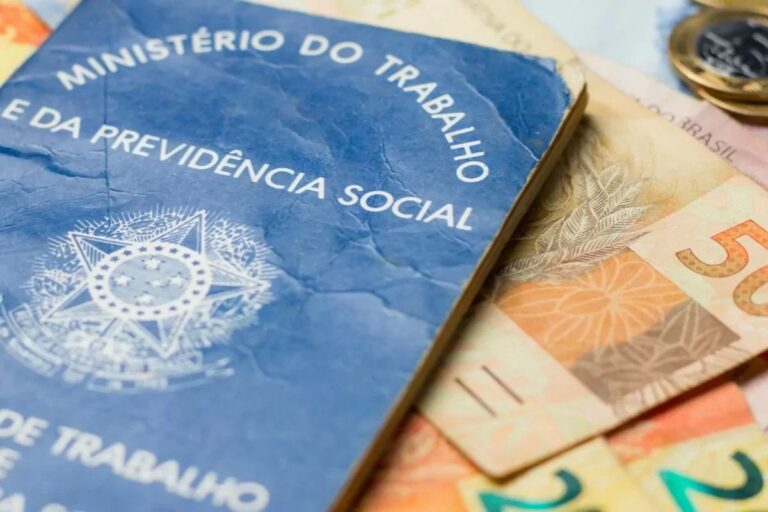 Uma Carteira de Trabalho e Previdência Social (CTPS) brasileira azul, em primeiro plano, sobreposta parcialmente a notas de Real (moeda brasileira) e moedas, simbolizando salário, emprego e direitos trabalhistas.