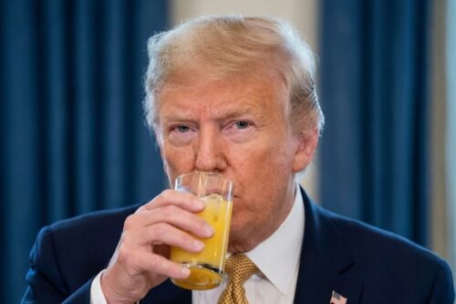 Donald Trump vestindo terno azul escuro e gravata amarela, segurando um copo de vidro com suco de laranja próximo à boca.