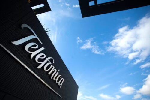 Perspectiva de baixo para cima mostrando o logotipo curvo e estilizado "Telefonica" em branco, fixado na fachada escura e moderna de um edifício corporativo. O céu está azul com algumas nuvens brancas, e a luz sugere um dia claro.