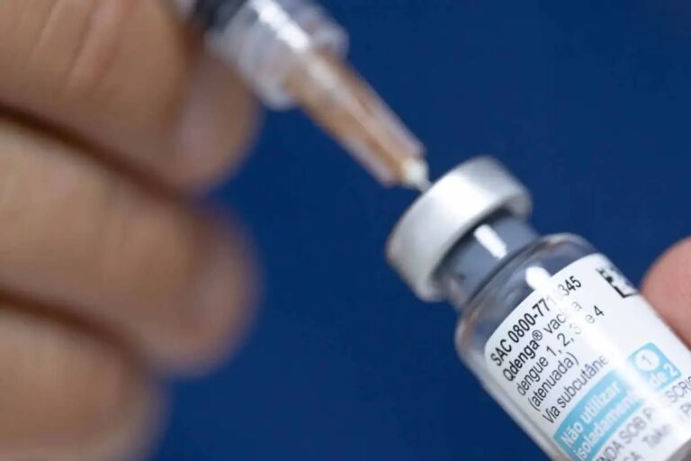 Close-up focado na tampa de um frasco da vacina contra a dengue, enquanto uma seringa tem sua agulha inserida no frasco para aspirar o líquido.