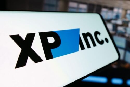 Close-up da tela de um dispositivo móvel exibindo o logotipo da empresa XP Inc. O logo é composto pelas letras 'XP' e 'inc.' na cor preta, separadas por uma forma geométrica azul. O fundo da imagem mostra um ambiente de escritório moderno e desfocado.