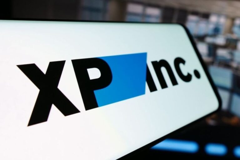 Close-up da tela de um dispositivo móvel exibindo o logotipo da empresa XP Inc. O logo é composto pelas letras 'XP' e 'inc.' na cor preta, separadas por uma forma geométrica azul. O fundo da imagem mostra um ambiente de escritório moderno e desfocado.