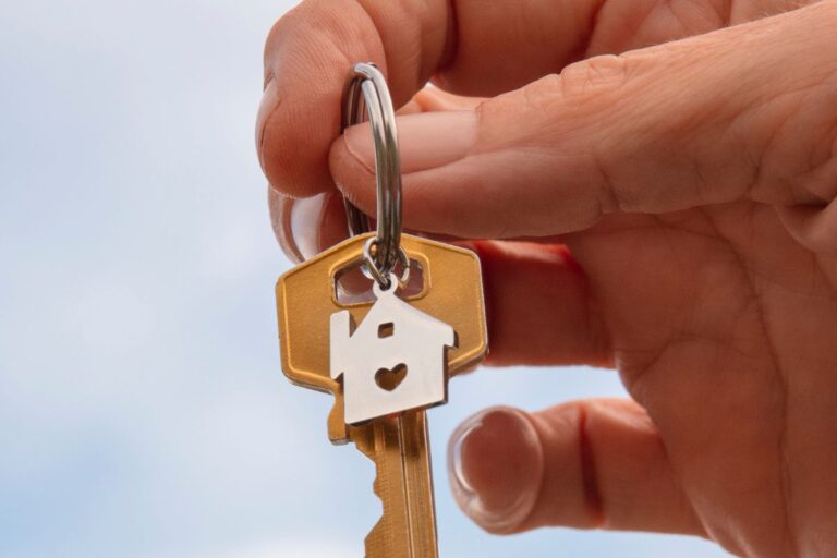 Close-up de uma mão segurando uma chave dourada, com um chaveiro metálico prateado em formato de casa e um pequeno coração vazado no centro.