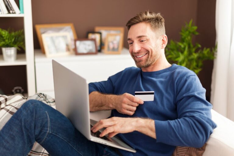 Homem sorridente sentado no sofá, usando um notebook para compras online e segurando um cartão de crédito.