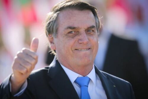 Close-up do político brasileiro Jair Bolsonaro, vestindo um terno escuro e gravata azul, sorrindo e fazendo um sinal de positivo com a mão direita levantada.