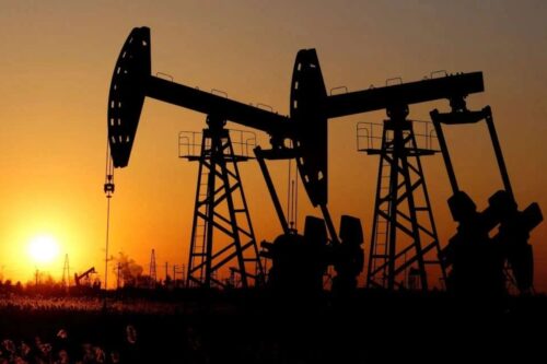 Silhueta de duas bombas de petróleo (cabeças de cavalo) em operação, com a imagem enquadrada por um pôr do sol laranja e avermelhado brilhante ao fundo.