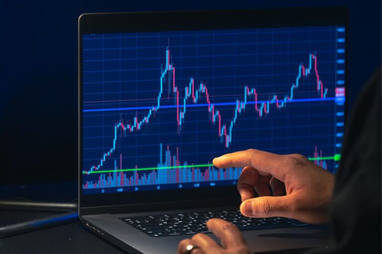 ma pessoa com um anel no dedo aponta para um gráfico de velas de trading (compra e venda de ativos) em ascensão em uma tela de notebook, no escuro.
