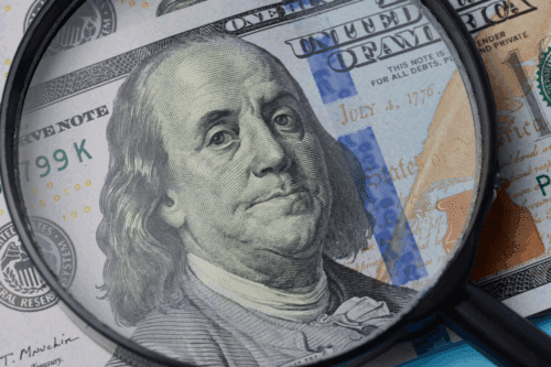 Exame detalhado de uma nota de dólar de cem dólares usando uma lupa, destacando a imagem de Benjamin Franklin, referência em dinheiro e investimento.