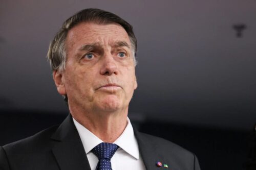 Fotografia em close do ex-presidente Jair Bolsonaro. Ele veste um paletó escuro, camisa branca e gravata azul com pequenos detalhes. Bolsonaro está com uma expressão séria, olhando levemente para cima e para o lado, contra um fundo neutro e desfocado.