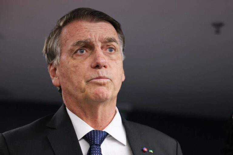 Fotografia em close do ex-presidente Jair Bolsonaro. Ele veste um paletó escuro, camisa branca e gravata azul com pequenos detalhes. Bolsonaro está com uma expressão séria, olhando levemente para cima e para o lado, contra um fundo neutro e desfocado.
