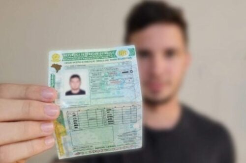 Mão segurando e exibindo uma Carteira Nacional de Habilitação (CNH) brasileira, dobrada ao meio, com o lado que contém a foto e os dados principais voltado para frente.